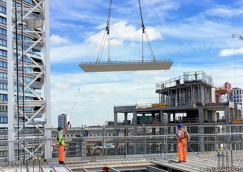 A precast concrete panel suspended from a crane over two workers on an open construction deck at height.
