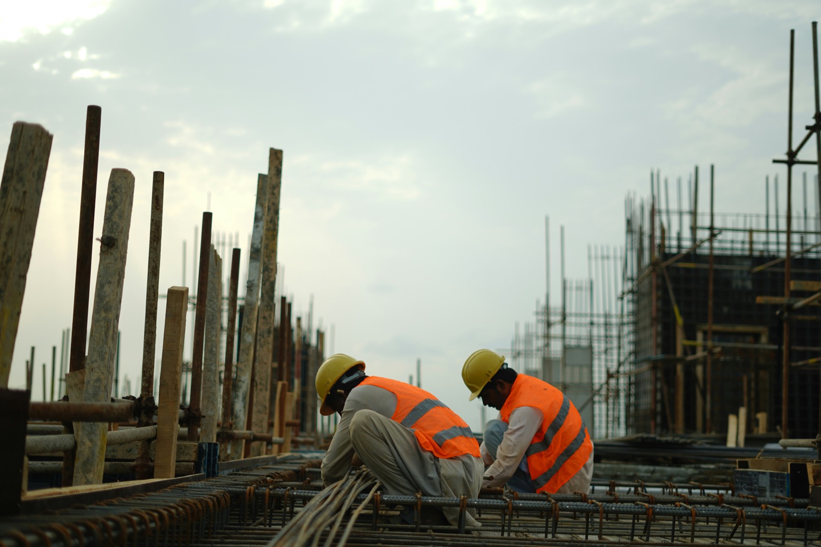 Two construction workers in high-visibility vests and hard hats tying rebar on a formwork deck, with scaffolding behind them